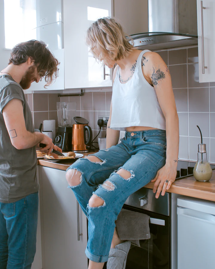 Pareja en la cocina: él corta pan, ella conversa sentada en el mesón